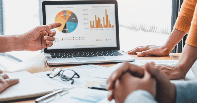 Equipe analisando gráficos de desempenho no notebook durante reunião estratégica, discutindo indicadores financeiros e margem de contribuição por categoria.