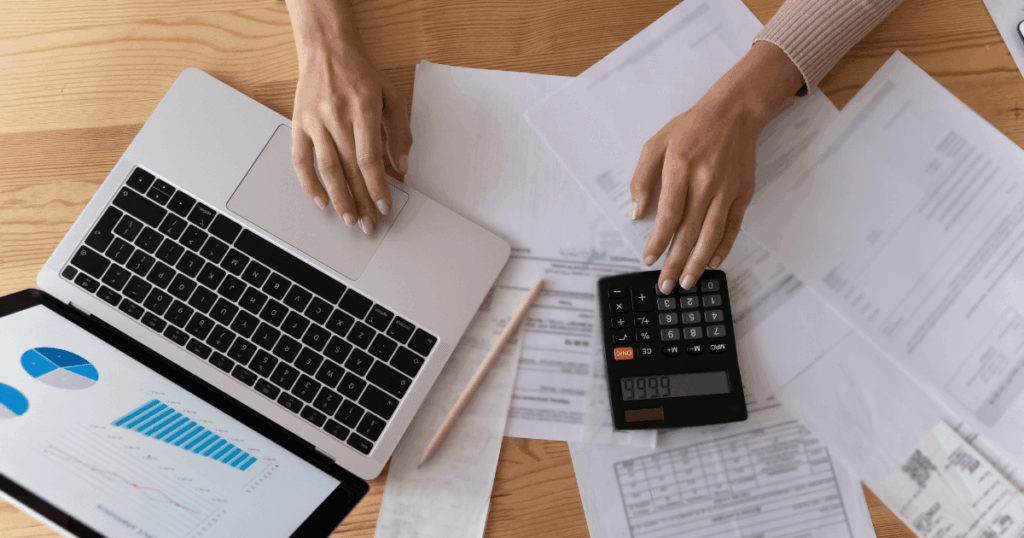 Pessoa analisando custos e despesas, com um laptop, uma calculadora e documentos financeiros espalhados sobre a mesa. Gráficos e relatórios são visíveis na cena.