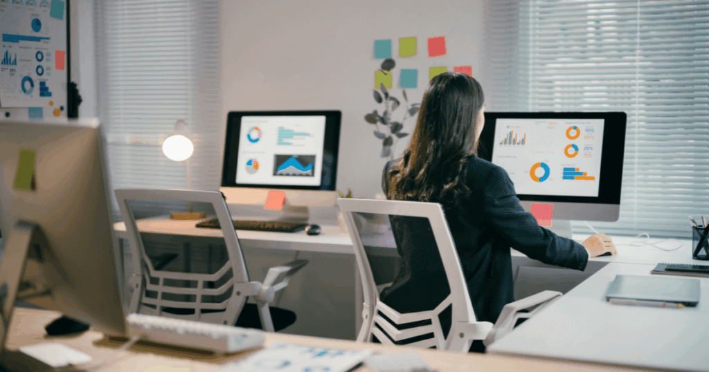 Profissional trabalhando em escritório moderno, analisando indicadores de gestão em dashboards com gráficos e métricas exibidos em telas de computador.