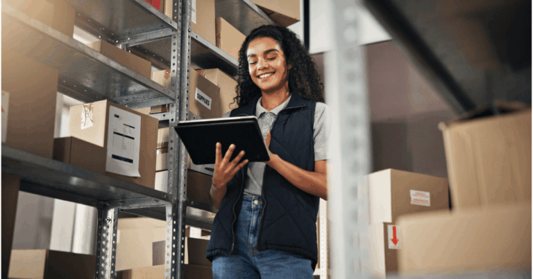Mulher sorrindo enquanto usa um tablet em um ambiente de estoque, com prateleiras de caixas ao fundo. A imagem ilustra a gestão de inventário, relacionada ao conceito de prazo médio de estocagem.