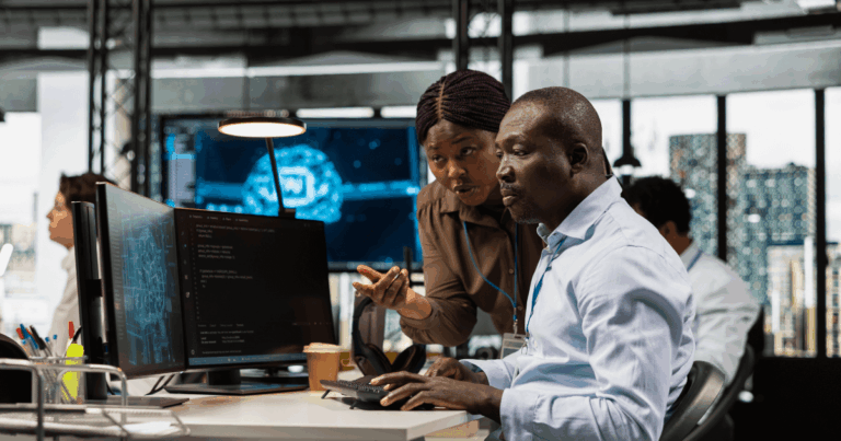 Profissionais de tecnologia analisando códigos e dados em monitores em um escritório moderno, representando gestão de TI e trabalho colaborativo na área de tecnologia da informação.