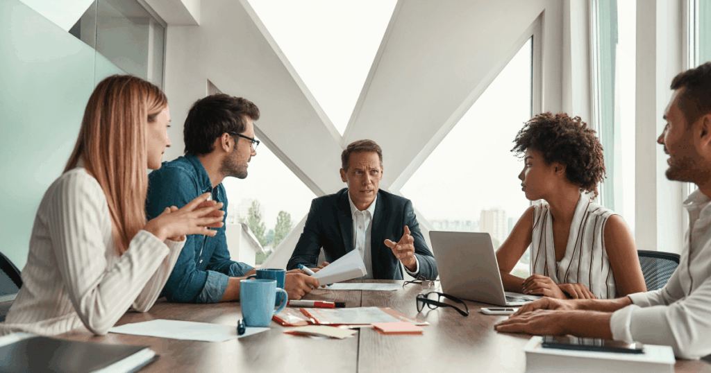Equipe executiva reunida em sala de reunião moderna discutindo estratégias de gestão empresarial com uso de relatórios e notebook.