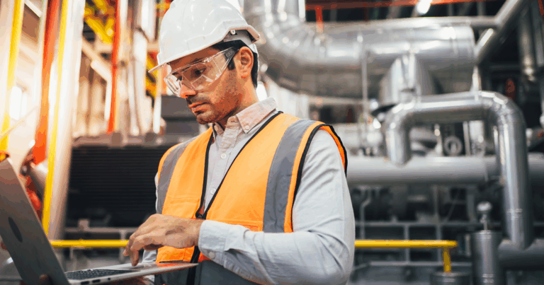 Profissional utilizando os equipamentos de EPi em uma indústria e analisando a gestão da qualidade em um notebook
