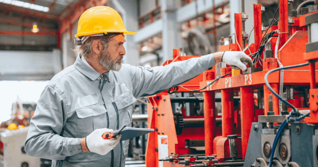 Técnico industrial utilizando capacete e tablet enquanto realiza inspeção em máquina na fábrica, representando a aplicação do MPS para padronização e melhoria de processos produtivos.