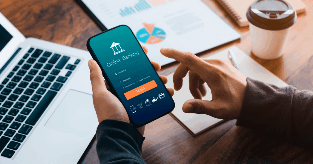 Pessoa acessando aplicativo de operações bancárias no celular, com gráficos financeiros e laptop sobre a mesa, representando gestão digital de contas e transações financeiras.