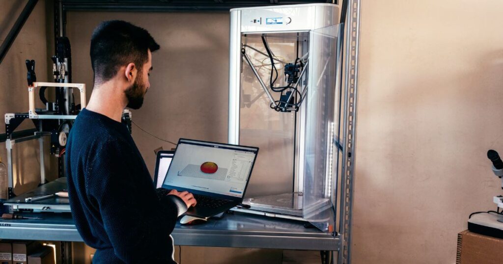 Profissional operando impressora 3D com modelagem digital em notebook, ilustrando aplicação prática da manufatura aditiva na produção de protótipos e peças industriais.