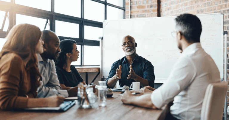Reunião estratégica para o crescimento empresarial, com um grupo diversificado de profissionais discutindo soluções de negócios enquanto um líder compartilha insights em frente a um quadro branco com planejamento e análise de estratégias.