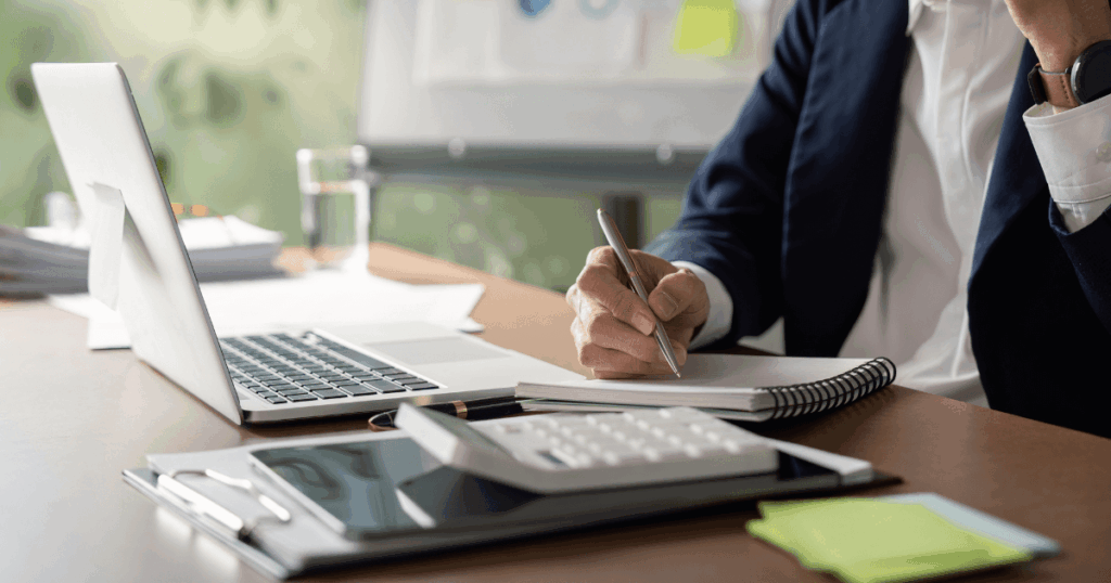 Pessoa escrevendo em um caderno, com laptop e calculadora em uma mesa, analisando tabela ICMS.