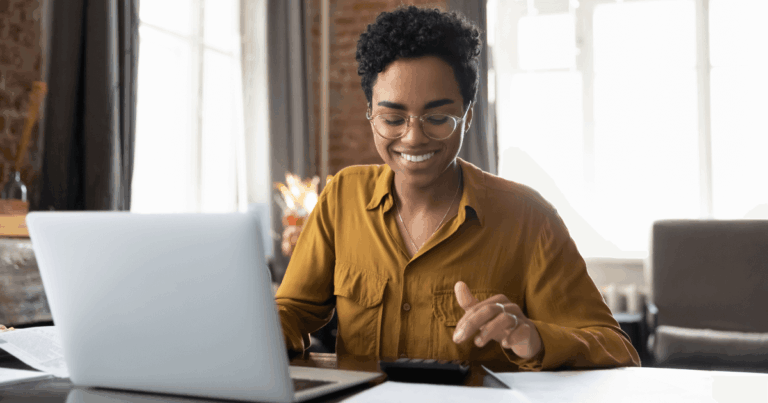 Mulher sorridente usando um laptop e calculadora, demonstrando prática de controle financeiro em um ambiente de trabalho organizado.