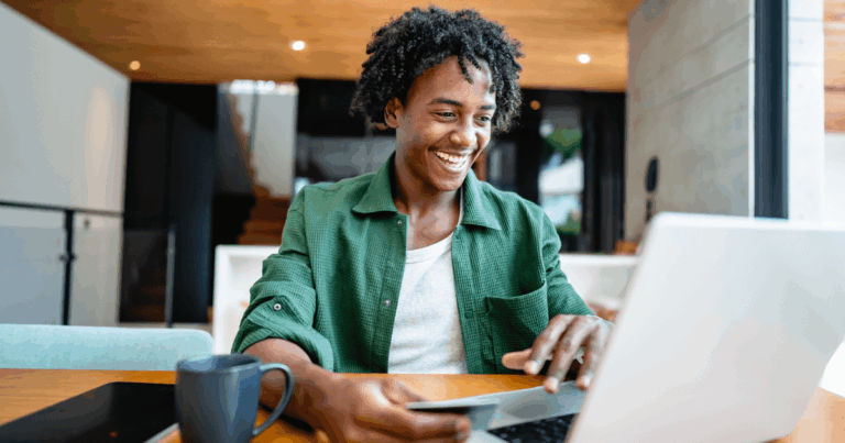 Homem sorridente utilizando um laptop e um cartão de crédito, ilustrando um processo de pagamento digital, possível referência ao conceito de Split Payment.