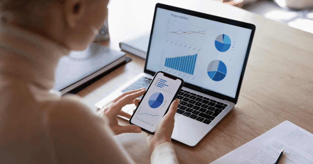 Profissional analisando gráficos sobre o capital de giro no notebook, com celular em mãos.