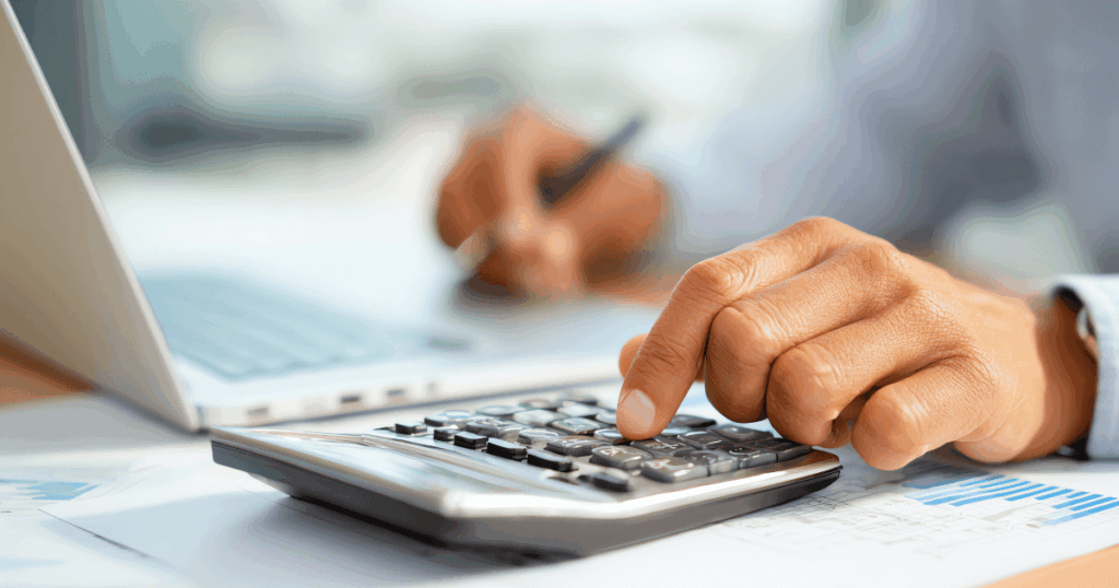 Pessoa utilizando uma calculadora e um laptop, com foco nas mãos, realizando cálculos financeiros relacionados a despesas fixas e variáveis.