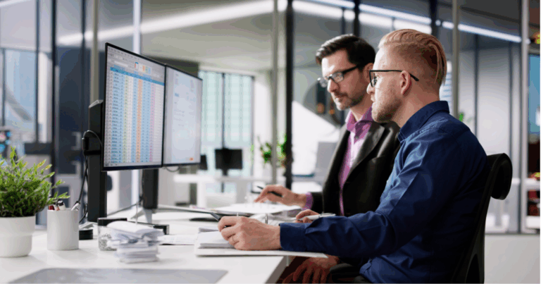 Dois profissionais analisando planilhas e dados financeiros em monitores duplos em um escritório moderno, representando processos de gestão fiscal e tributária eficientes.
