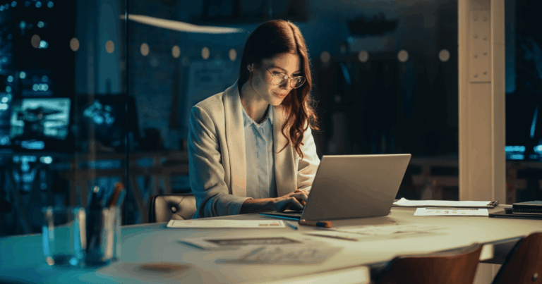 Mulher trabalhando em um escritório à noite diante do laptop, analisando documentos e realizando atividades relacionadas ao adicional noturno.