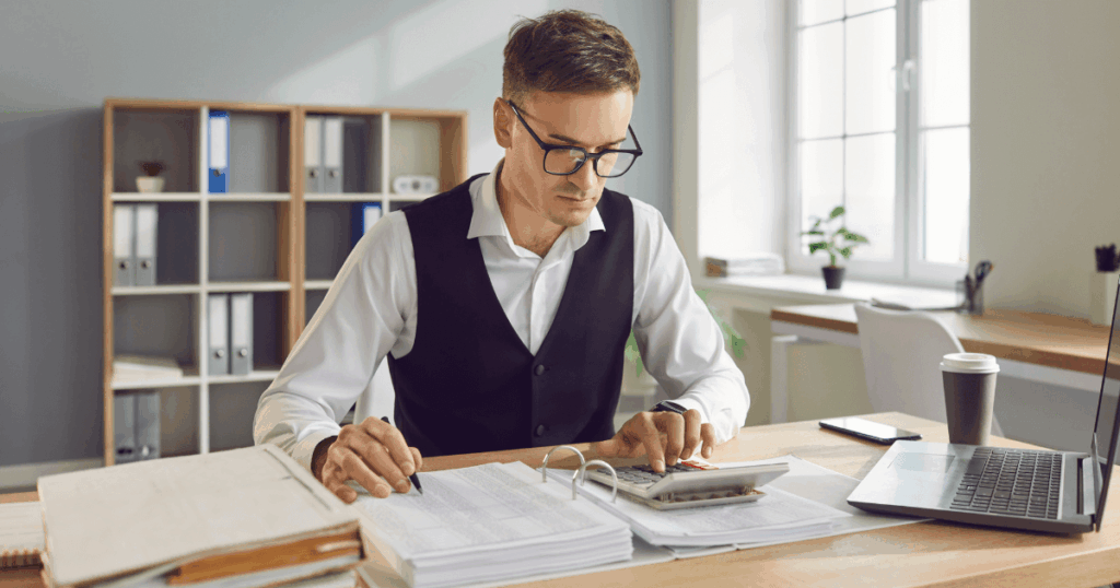 Homem em um escritório usando calculadora e analisando documentos para calcular horas trabalhadas, sentado à mesa com laptop, papéis e pastas ao fundo.