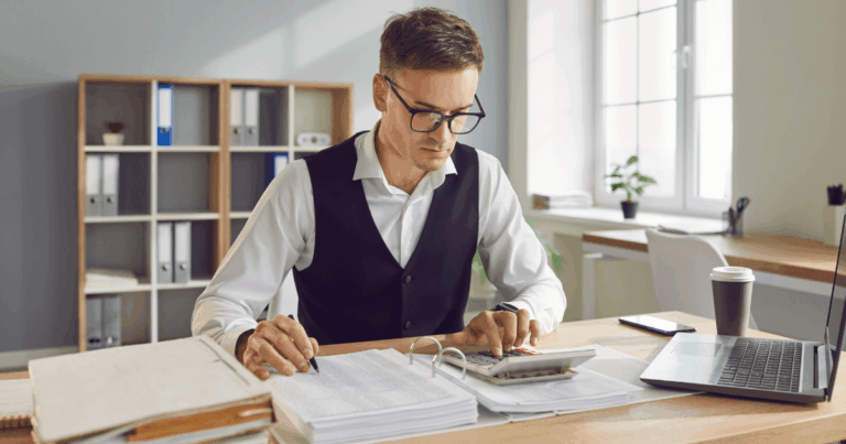 Homem em um escritório usando calculadora e analisando documentos para calcular horas trabalhadas, sentado à mesa com laptop, papéis e pastas ao fundo.