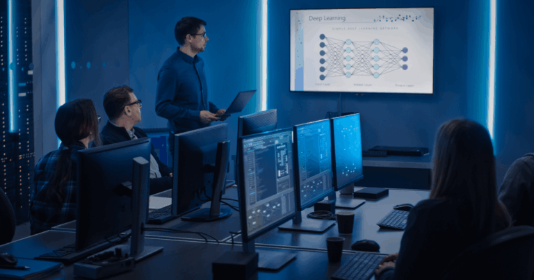Equipe em uma sala de TI observando uma apresentação sobre deep learning, com um instrutor explicando uma rede neural exibida em um telão e vários computadores ligados à frente dos participantes.