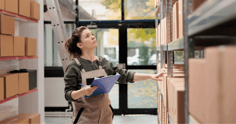 Profissional em um estoque verificando caixas nas prateleiras com prancheta na mão, representando o processo de entrega e controle de pedidos.