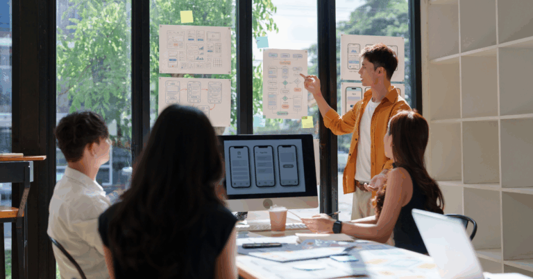 quipe reunida em uma sala de reunião colaborativa, analisando protótipos de telas e fluxos de aplicativo em um quadro de vidro, discutindo soluções digitais relacionadas a benefícios corporativos.