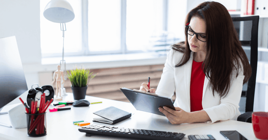 Profissional trabalhando em escritório, anotando informações financeiras em uma prancheta sobre a mesa, representando atividades de tesouraria e gestão financeira.