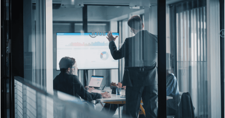 Equipe em reunião corporativa analisando gráficos de desempenho em uma tela, discutindo estratégias de vendas sazonais em um escritório moderno com paredes de vidro.