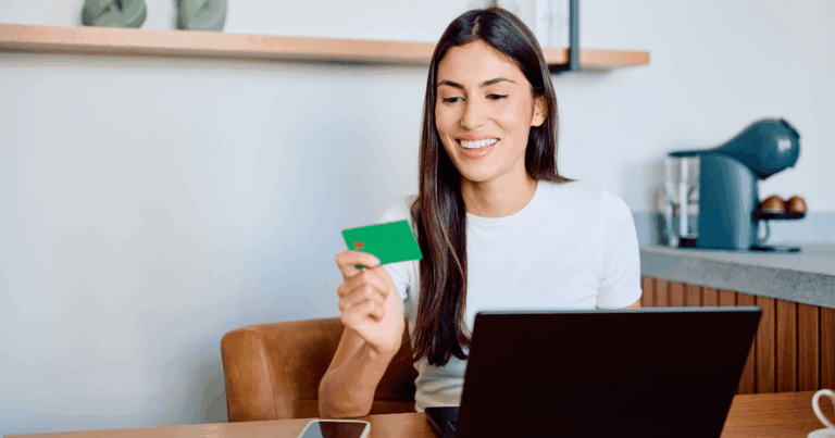 Mulher sorrindo enquanto segura um cartão de crédito e usa o notebook para fazer uma compra online, representando o comportamento do consumidor no ambiente digital.