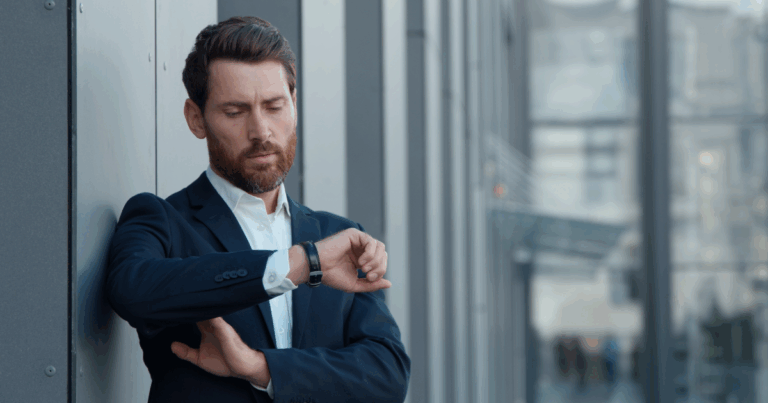 Homem de terno encostado em um prédio corporativo olhando para o relógio de pulso com expressão preocupada, representando atraso no trabalho em ambiente urbano moderno.