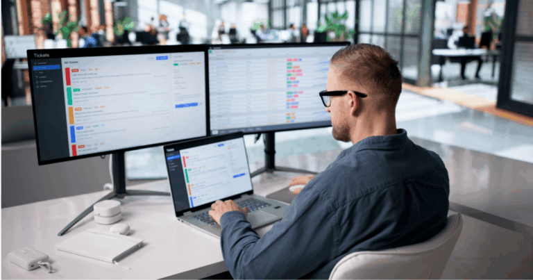 Profissional trabalhando em estação com múltiplos monitores e notebook, acompanhando painéis e listas de informações em tempo real. A cena representa processos de integração de dados.
