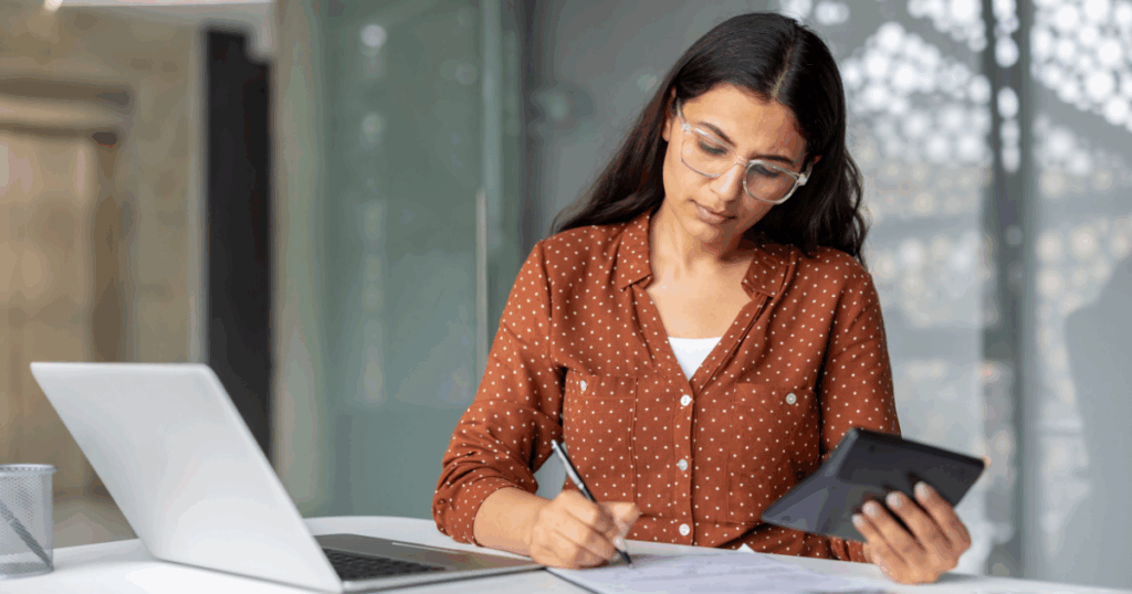 Mulher analisando documentos e fazendo anotações enquanto usa um tablet e um notebook, representando atividades de cálculo e gestão tributária relacionadas ao DIFAL